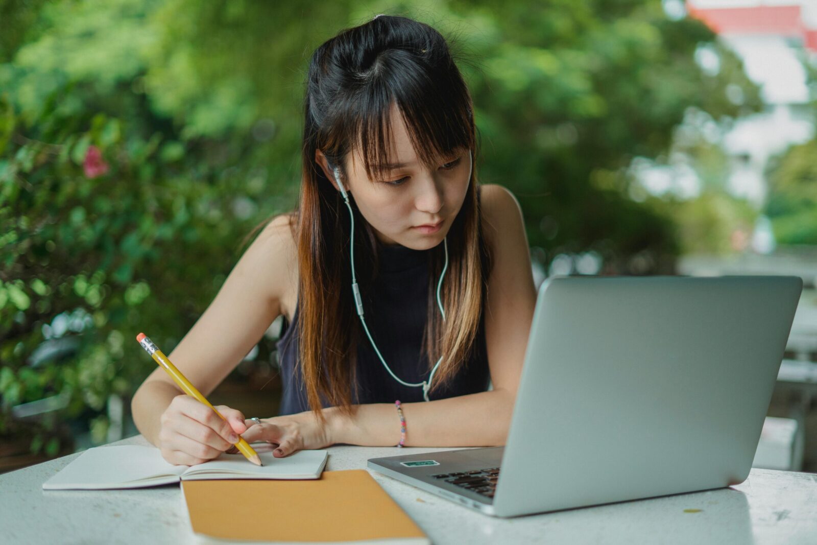 Concentrated Asian female in black blouse watching video lecture at laptop and taking notes sitting at table in cafe