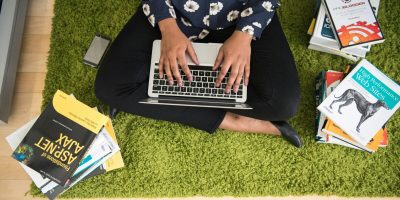 A woman sits on a green carpet working on a laptop with stacks of books around her, focusing on technology projects.