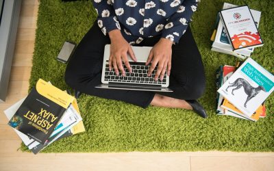 A woman sits on a green carpet working on a laptop with stacks of books around her, focusing on technology projects.