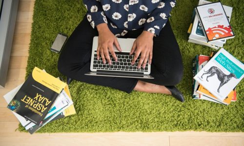 A woman sits on a green carpet working on a laptop with stacks of books around her, focusing on technology projects.