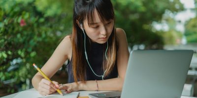 Concentrated Asian female in black blouse watching video lecture at laptop and taking notes sitting at table in cafe