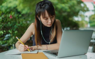 Concentrated Asian female in black blouse watching video lecture at laptop and taking notes sitting at table in cafe